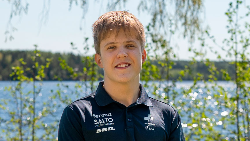 Aaron Hänninen hymyilemässä kameralle auringossa. Taustalla vihreää kasvustoa ja vesistö.